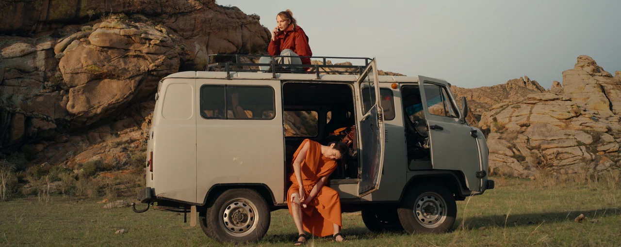 A woman sits on the roof of a UAZ van among the rocks; a monk sits in its open doorway.